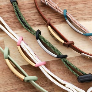 May include: Close-up of several colorful bracelets with various designs. The bracelets feature braided cords in white, green, and brown, accented with gold, silver, and black metal bars. Some have pink and green accents.