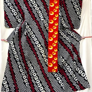 Puede incluir: Un kimono japonés tradicional con un patrón llamativo. La prenda presenta un diseño repetitivo de cuadrados blancos sobre un fondo negro, intercalados con rayas rojas. Una faja roja con diseños circulares amarillos añade un contraste vibrante.