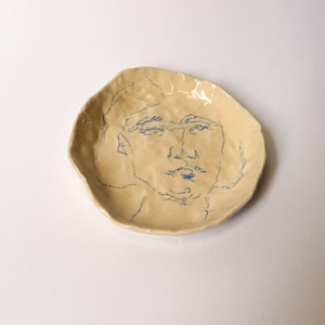 Puede incluir: Un plato de cerámica de color crema, de forma redonda con un borde irregular y ondulado. La superficie presenta una ilustración de líneas azules dibujada a mano de un rostro humano en el centro. El material cerámico tiene un acabado brillante.