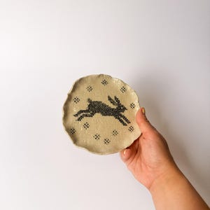 May include: A small, round, off-white ceramic dish with a wavy, uneven edge. The surface features a textured, dotted pattern and a central black graphic illustration of a leaping rabbit, surrounded by small, black, cross-shaped decorative accents.