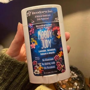 May include: A white deodorant stick with the text "Deodorwise" and "MOODY JUDY" in a floral design. The label lists "Lavender, Geranium, Balsam & Amyris" and states "No Aluminum, No Baking soda, No Parabens". The product is 2.65 oz / 75 ml.