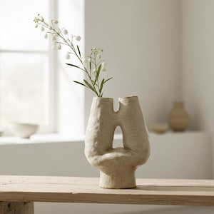 Może przedstawiać: Beżowy, teksturowany ceramiczny wazon o unikalnym, abstrakcyjnym kształcie. Wazon ma dwa otwory u góry i duży otwór pośrodku. Zawiera gałązkę białych kwiatów z zielonymi liśćmi, umieszczoną na drewnianej powierzchni.