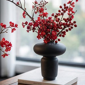 以下が含まれることがあります： ユニークな丸みを帯びた上部を持つ黒いセラミック花瓶には、鮮やかな赤い花とベリーが入っています。花瓶は白い本の上に置かれ、背景には追加の赤い花があります。花瓶はモダンでミニマリストなデザインです。