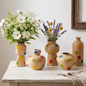 Puede incluir: Una colección de jarrones de cerámica beige con lunares coloridos. Los jarrones, de diversas formas y tamaños, contienen flores frescas. El arreglo incluye margaritas, lavanda y flores amarillas globulares, creando una exhibición vibrante y alegre.