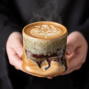 May include: A handmade ceramic mug with a distinctive, textured glaze in brown, green, and red hues. The mug contains a latte, adorned with detailed latte art. Steam rises from the warm drink, suggesting a comforting beverage.