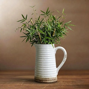 Può includere: Una brocca in ceramica bianca con un design a coste, contenente steli e foglie di bambù verdi. La brocca ha un manico con un motivo decorativo e una base marrone. Lo sfondo è marrone caldo.