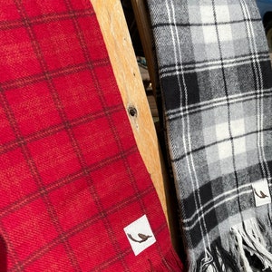 May include: Two plaid blankets hanging on a wooden chair. The blanket on the left is red with black plaid, and the blanket on the right is black and white plaid. Both blankets have a small bird logo tag.