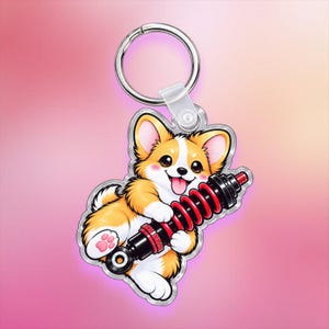 Peut inclure: Un porte-clés en acrylique transparent représentant une illustration de dessin animé d'un chien Corgi beige et blanc tenant un amortisseur de suspension automobile noir et rouge. Il comprend un anneau en métal argenté et un connecteur en plastique.
