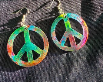 May include: Two colorful peace sign earrings. Each earring features a peace symbol in a circular shape with a hook for the ear. The earrings are a mix of green, blue, orange, and pink colors.