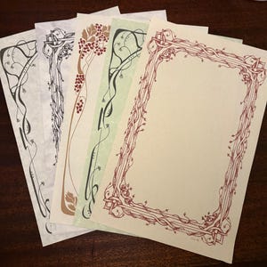 May include: A collection of decorative stationery sheets with Art Nouveau-style borders. The designs feature flowing lines, vines, and floral elements in various colors, including black, brown, and red. The paper has a cream color.