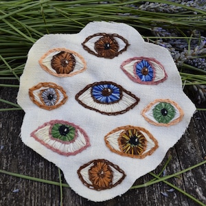 May include: A white fabric with nine embroidered eyes in different colors, including blue, green, brown, and orange. The eyes are surrounded by a white fabric with a slightly textured surface.
