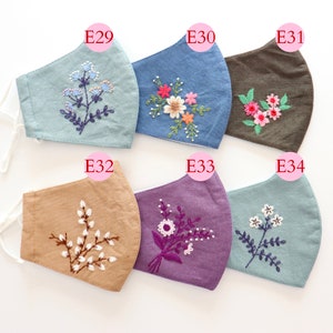 May include: Six different face masks with embroidered floral designs. The masks are made of various fabrics in colours like blue, brown, beige, purple, and light blue. Each mask has a unique floral design embroidered on it.
