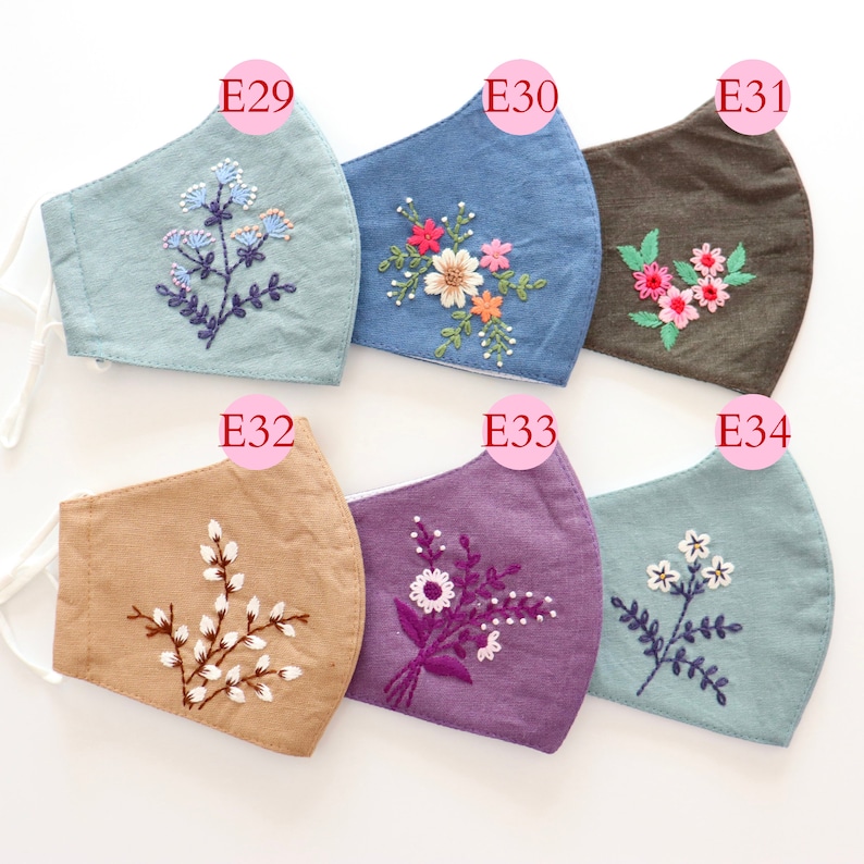 May include: Six different face masks with embroidered floral designs. The masks are made of various fabrics in colours like blue, brown, beige, purple, and light blue. Each mask has a unique floral design embroidered on it.