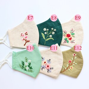 May include: Six different fabric face masks with embroidered floral designs. The masks are in various colours, including beige, green, and olive green. The designs include a small branch of pink flowers, a sprig of white flowers, a cluster of red flowers, a bouquet of pink and purple flowers, and a branch of yellow flowers.