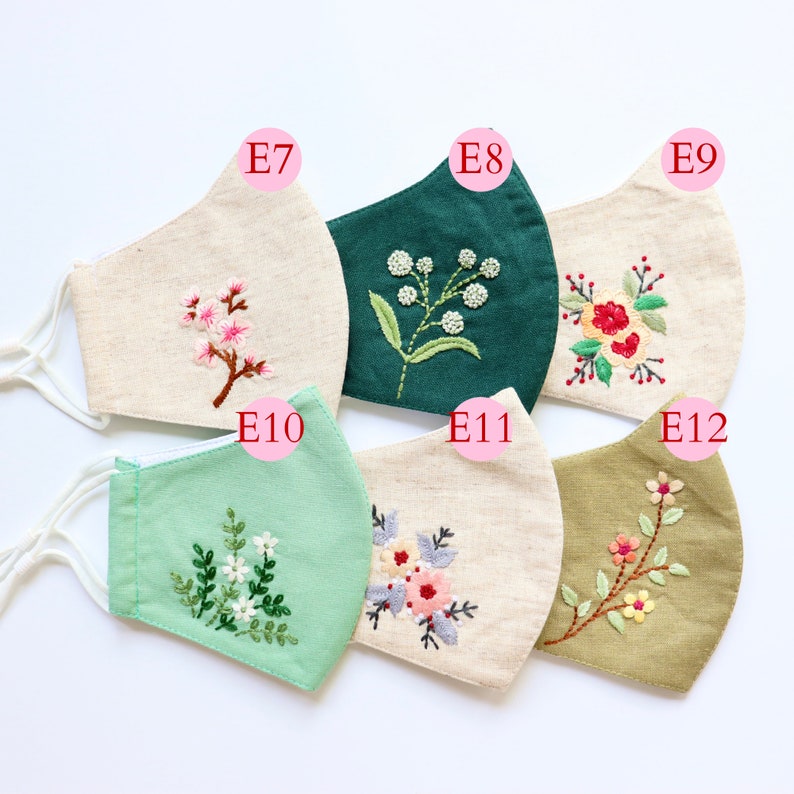 May include: Six different fabric face masks with embroidered floral designs. The masks are in various colours, including beige, green, and olive green. The designs include a small branch of pink flowers, a sprig of white flowers, a cluster of red flowers, a bouquet of pink and purple flowers, and a branch of yellow flowers.