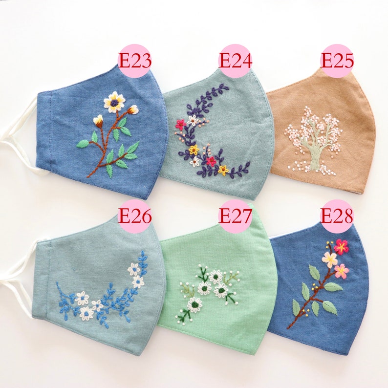 May include: Six different face masks with embroidered floral designs. The masks are made of a light blue, light green, and beige fabric. The designs include a tree, a wreath, and various flowers.