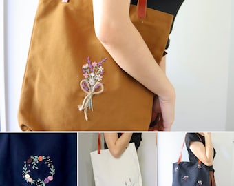 May include: Three tote bags with different embroidered floral designs. The first bag is brown with a bouquet of purple, pink, and white flowers. The second bag is white with a small bouquet of purple flowers. The third bag is black with a small bouquet of pink and white flowers.