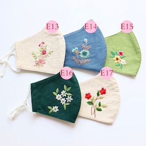 May include: Five different fabric face masks with embroidered floral designs. The masks are in various colours, including beige, green, blue, and dark green. The masks are labelled with numbers E13, E14, E15, E16, and E17.