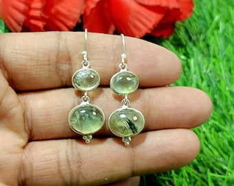 May include: A pair of silver drop earrings featuring two oval-shaped, light green translucent stones per earring. Each stone contains visible dark needle-like inclusions. The earrings hang from silver hooks and have small decorative silver beads at the base.