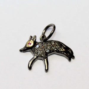 Puede incluir: Colgante de zorro de metal oscuro con un lazo para una cadena. El zorro tiene un ojo de piedra preciosa roja y está incrustado con pequeñas piedras brillantes. El colgante está sobre un fondo blanco.