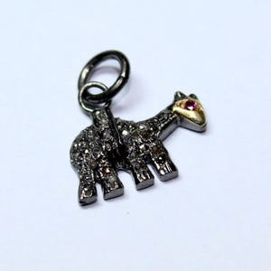 May include: A small, detailed charm in the shape of a giraffe. The charm is covered in small, sparkling stones, with a gold-colored face and a small, red gemstone eye. The charm has a dark metal finish and a loop for attaching to a necklace or bracelet.