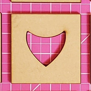 May include: A square wooden cutout with a heart-shaped design. The cutout is light brown and sits on a pink grid background. The heart shape is cut out of the wood, revealing the pink grid beneath.