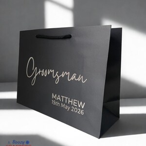 May include: A black gift bag with the word "Groomsman" in elegant script. Below, it reads "MATTHEW 15th May 2026". The bag has a black rope handle and is designed for a special occasion.