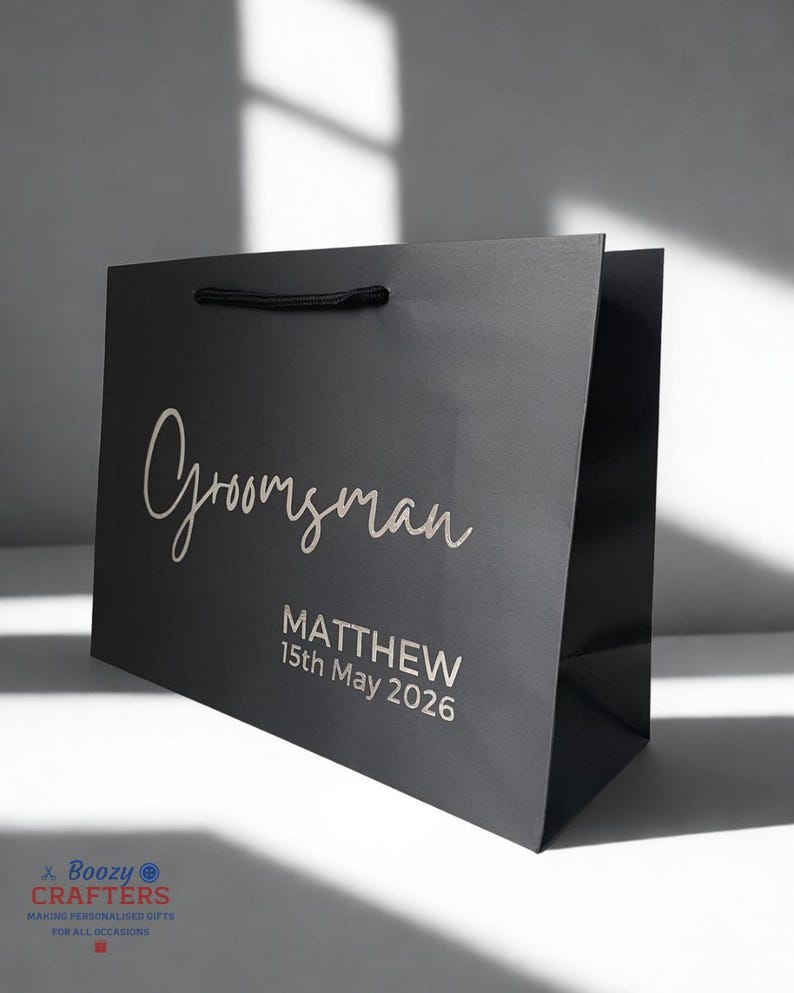 May include: A black gift bag with the word "Groomsman" in elegant script. Below, it reads "MATTHEW 15th May 2026". The bag has a black rope handle and is designed for a special occasion.