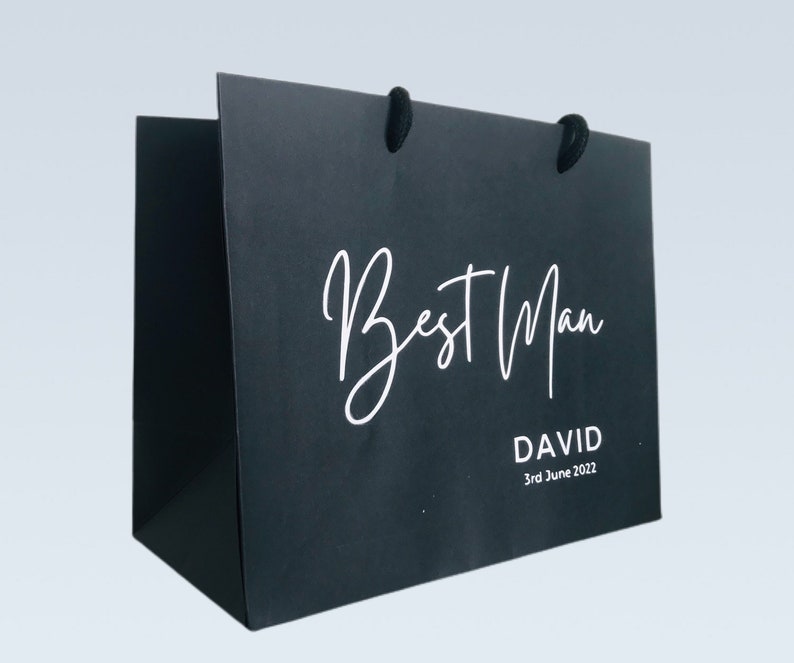 May include: Black gift bag with the words "Best Man" in white script. Below the text is the name "DAVID" and the date "3rd June 2022" in a smaller, white font. The bag has black handles.