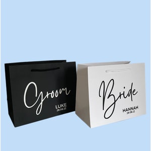 May include: Two gift bags, one black and one white, with the words "Groom" and "Bride" in elegant script. The black bag has "Luke 28.08.22" and the white bag has "Hannah 28.08.22". Both bags have matching rope handles.