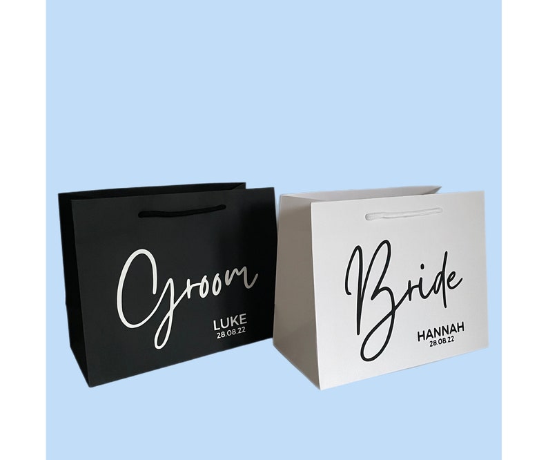 May include: Two gift bags, one black and one white, with the words "Groom" and "Bride" in elegant script. The black bag has "Luke 28.08.22" and the white bag has "Hannah 28.08.22". Both bags have matching rope handles.