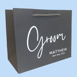 May include: A dark gray gift bag with the word "Groom" in white script. Below the word "Groom" is the name "MATTHEW" and the date "28th May 2022" in a smaller font. The bag has a gray rope handle.