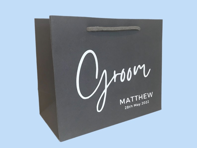 May include: A dark gray gift bag with the word "Groom" in white script. Below the word "Groom" is the name "MATTHEW" and the date "28th May 2022" in a smaller font. The bag has a gray rope handle.