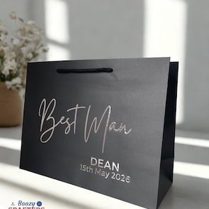 May include: A black gift bag with the words "Best Man" in silver script. Below, the name "DEAN" and the date "15th May 2026" are printed. The bag has a black cord handle and is likely for a wedding.