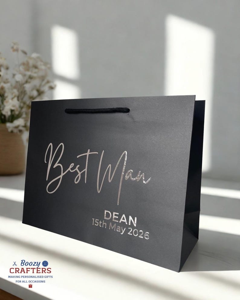 May include: A black gift bag with the words "Best Man" in silver script. Below, the name "DEAN" and the date "15th May 2026" are printed. The bag has a black cord handle and is likely for a wedding.