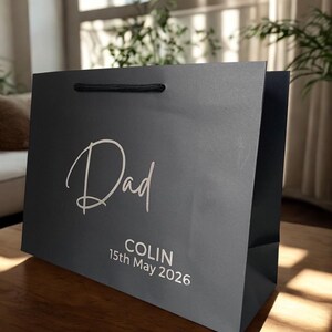 May include: A dark gray gift bag with the word "Dad" in elegant white script. Below, the name "COLIN" and the date "15th May 2026" are printed in white. The bag has a black rope handle.