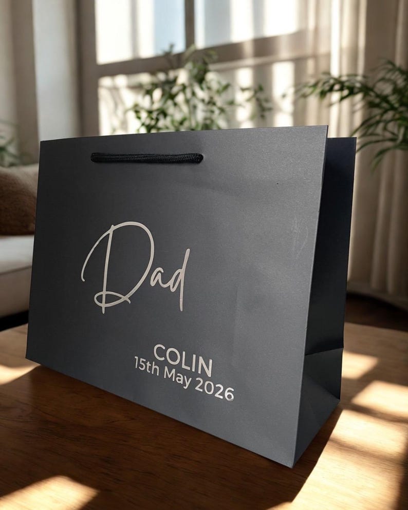 May include: A dark gray gift bag with the word "Dad" in elegant white script. Below, the name "COLIN" and the date "15th May 2026" are printed in white. The bag has a black rope handle.
