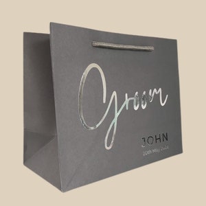May include: Gray gift bag with the word "Groom" in silver script. Below the word "Groom" is the name "JOHN" and the date "20th May 2022". The bag has a silver handle.