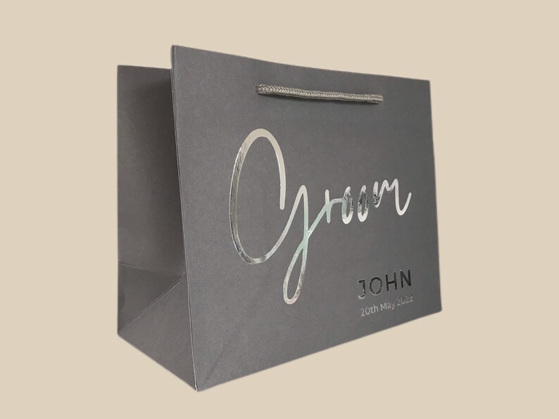 May include: Gray gift bag with the word "Groom" in silver script. Below the word "Groom" is the name "JOHN" and the date "20th May 2022". The bag has a silver handle.