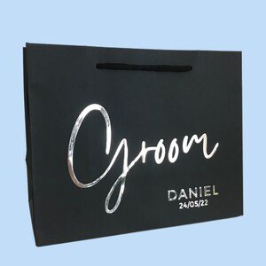 May include: Black gift bag with the word "Groom" in silver script. The name "Daniel" and the date "24/05/22" are printed below. The bag has a black cord handle and is set against a light blue background.