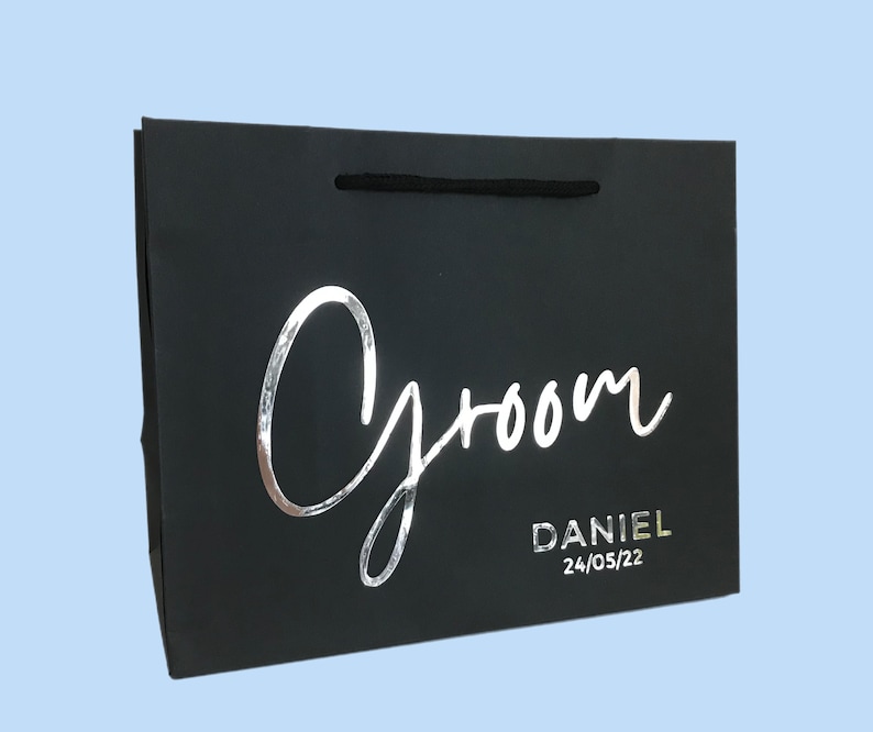 May include: Black gift bag with the word "Groom" in silver script. The name "Daniel" and the date "24/05/22" are printed below. The bag has a black cord handle and is set against a light blue background.