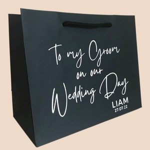 May include: Dark gray gift bag with black rope handles. White script reads "To my Groom on our Wedding Day" with the name "Liam" and the date "27.07.22" below. A wedding-themed gift bag.