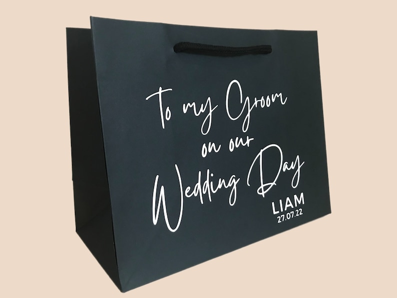 May include: Dark gray gift bag with black rope handles. White script reads "To my Groom on our Wedding Day" with the name "Liam" and the date "27.07.22" below. A wedding-themed gift bag.