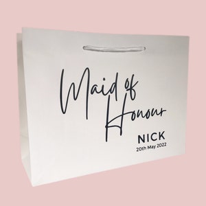 May include: White gift bag with the words "Maid of Honour" in elegant black script. Below the text is the name "NICK" and the date "20th May 2022". The bag has a white ribbon handle.