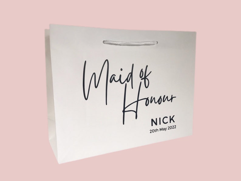 May include: White gift bag with the words "Maid of Honour" in elegant black script. Below the text is the name "NICK" and the date "20th May 2022". The bag has a white ribbon handle.
