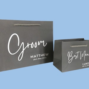 May include: Two gray gift bags with white script text. The larger bag says "Groom" and "Matthew 28th May 2022". The smaller bag says "Best Man" and "Luke 28th May 2022". Both bags have gray rope handles.