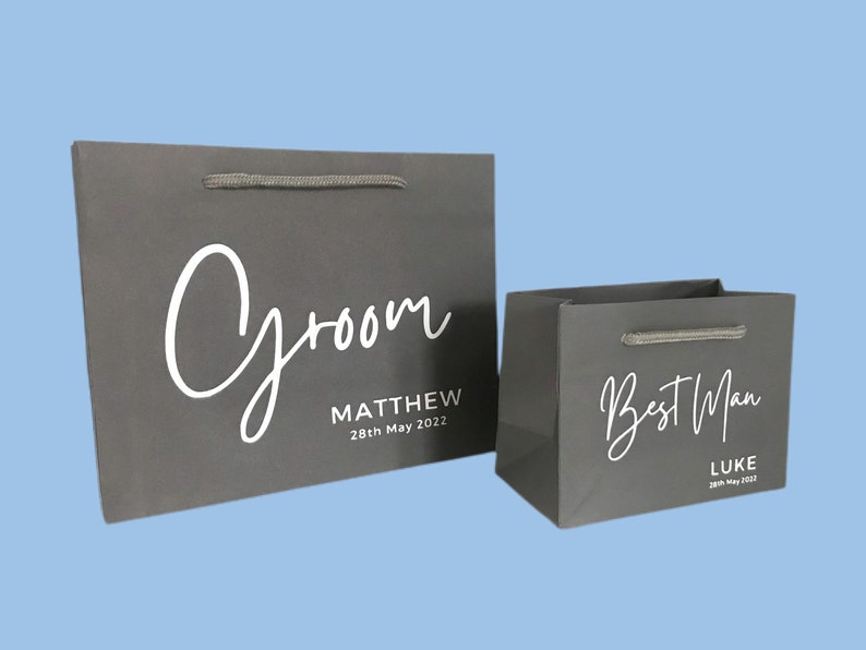 May include: Two gray gift bags with white script text. The larger bag says "Groom" and "Matthew 28th May 2022". The smaller bag says "Best Man" and "Luke 28th May 2022". Both bags have gray rope handles.