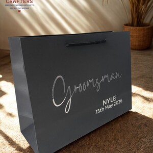 May include: A dark gray gift bag with the word "Groomsman" in silver script. The name "Nyle" and the date "15th May 2026" are printed below. The bag has black rope handles and the "Boozy Crafters" logo.