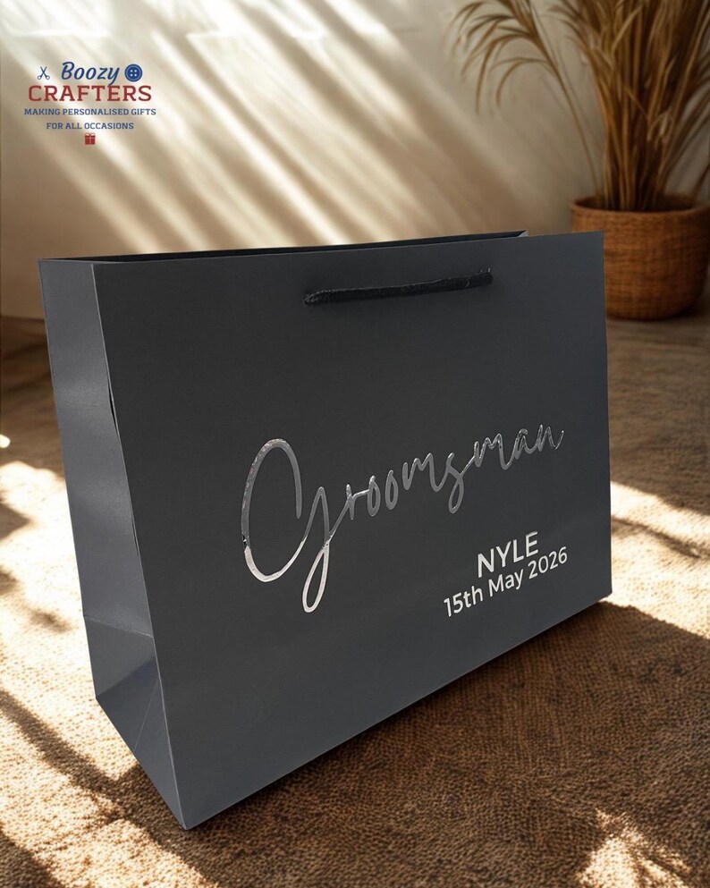 May include: A dark gray gift bag with the word "Groomsman" in silver script. The name "Nyle" and the date "15th May 2026" are printed below. The bag has black rope handles and the "Boozy Crafters" logo.