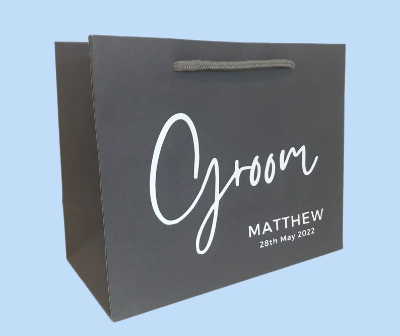 May include: A dark gray gift bag with the word "Groom" in white script. Below the word "Groom" is the name "MATTHEW" and the date "28th May 2022" in white text. The bag has a gray rope handle.